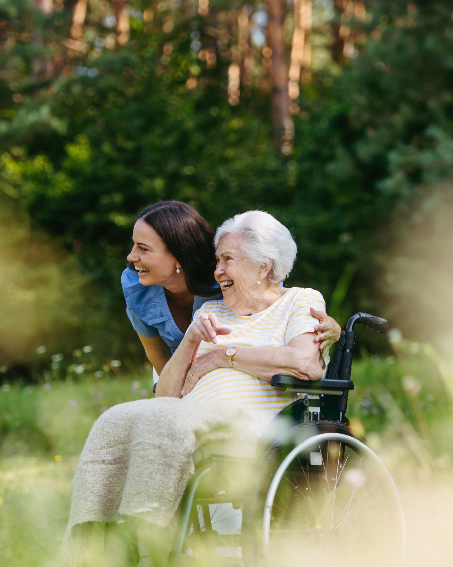 A caregiver leaning in beside a senior in a wheelchair, both smiling outdoors in a meadow.